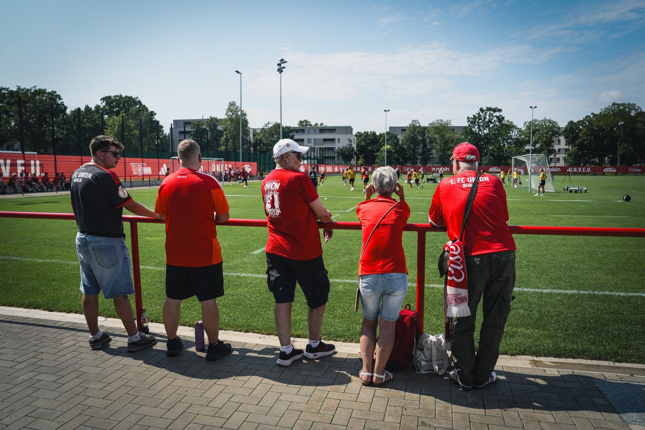 Fünf Personen in roten T-Shirts beobachten ein Fußballtraining auf einem grünen Platz.