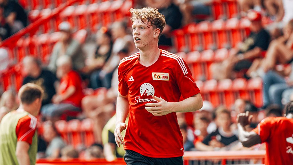 A young soccer player in a red jersey runs on the field, with spectators in the stands in the background.