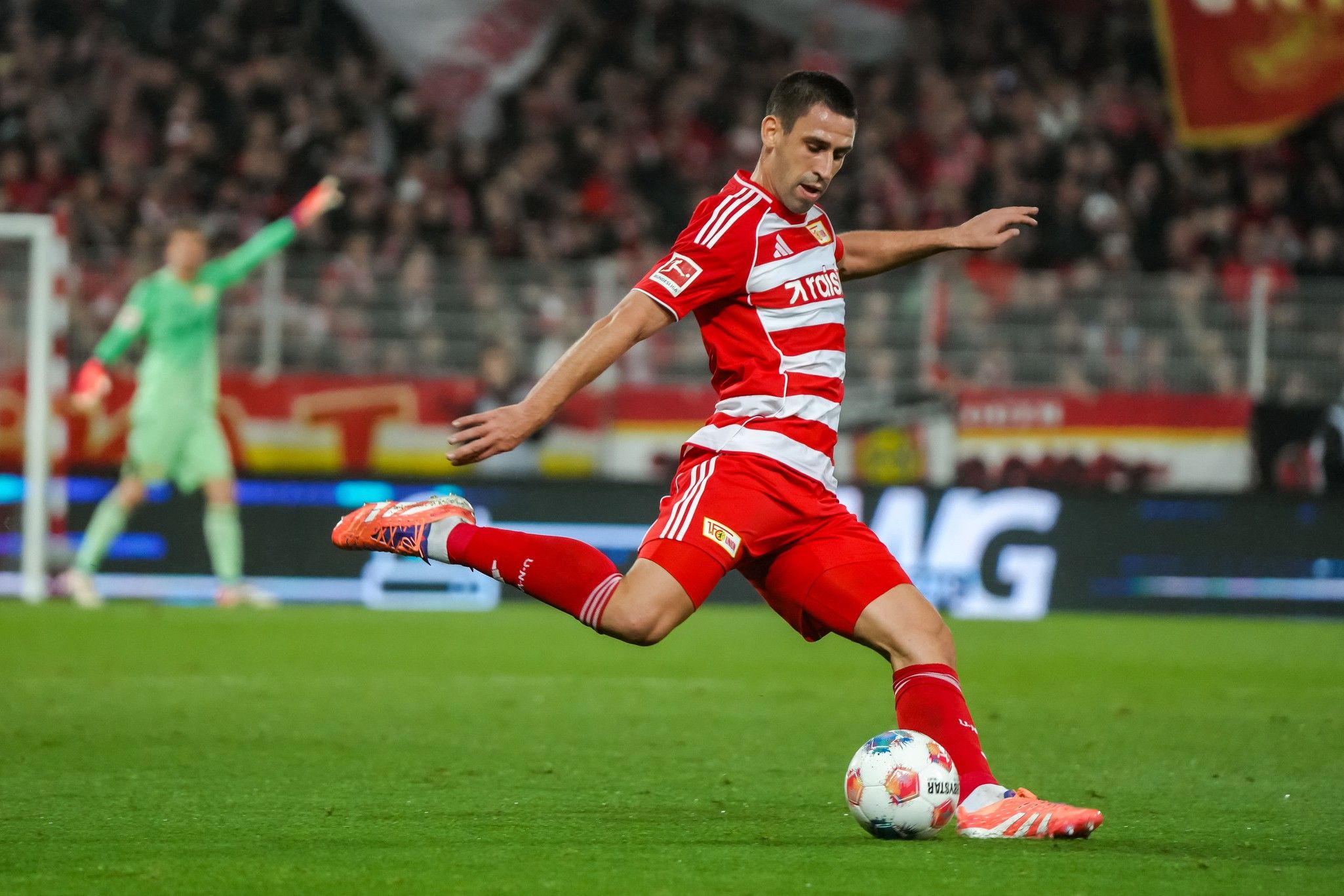 Spieler in rotem Fußballtrikot tritt den Ball auf dem Spielfeld, im Hintergrund jubelnde Fans und ein Torwart.