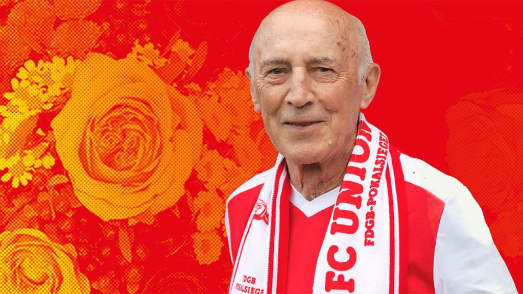 Älterer Mann mit Glatze, trägt ein rotes und weißes Fußballtrikot sowie einen Schal, vor einem roten Hintergrund mit Blumenmotiv.