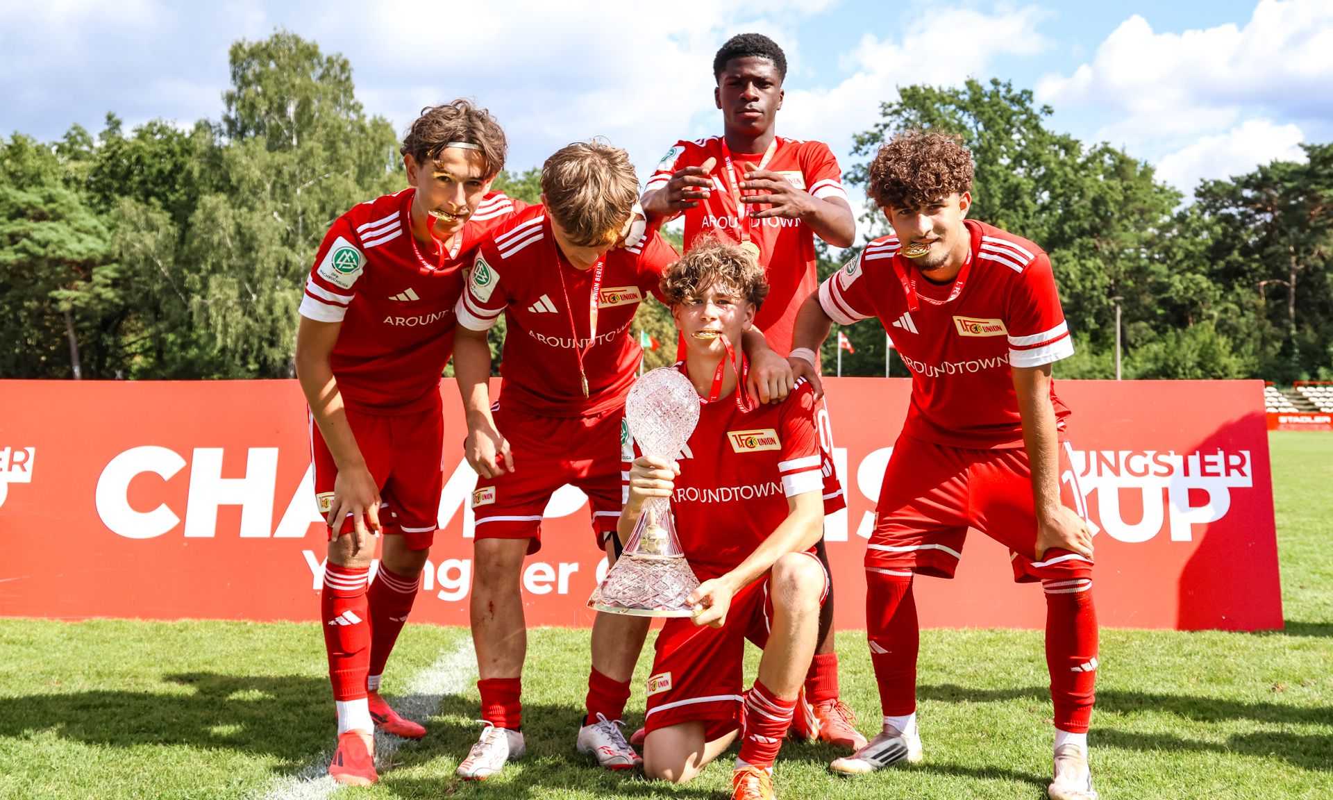 Fünf junge Fußballspieler in roten Trikots posieren mit einem Pokal auf einem Sportplatz.