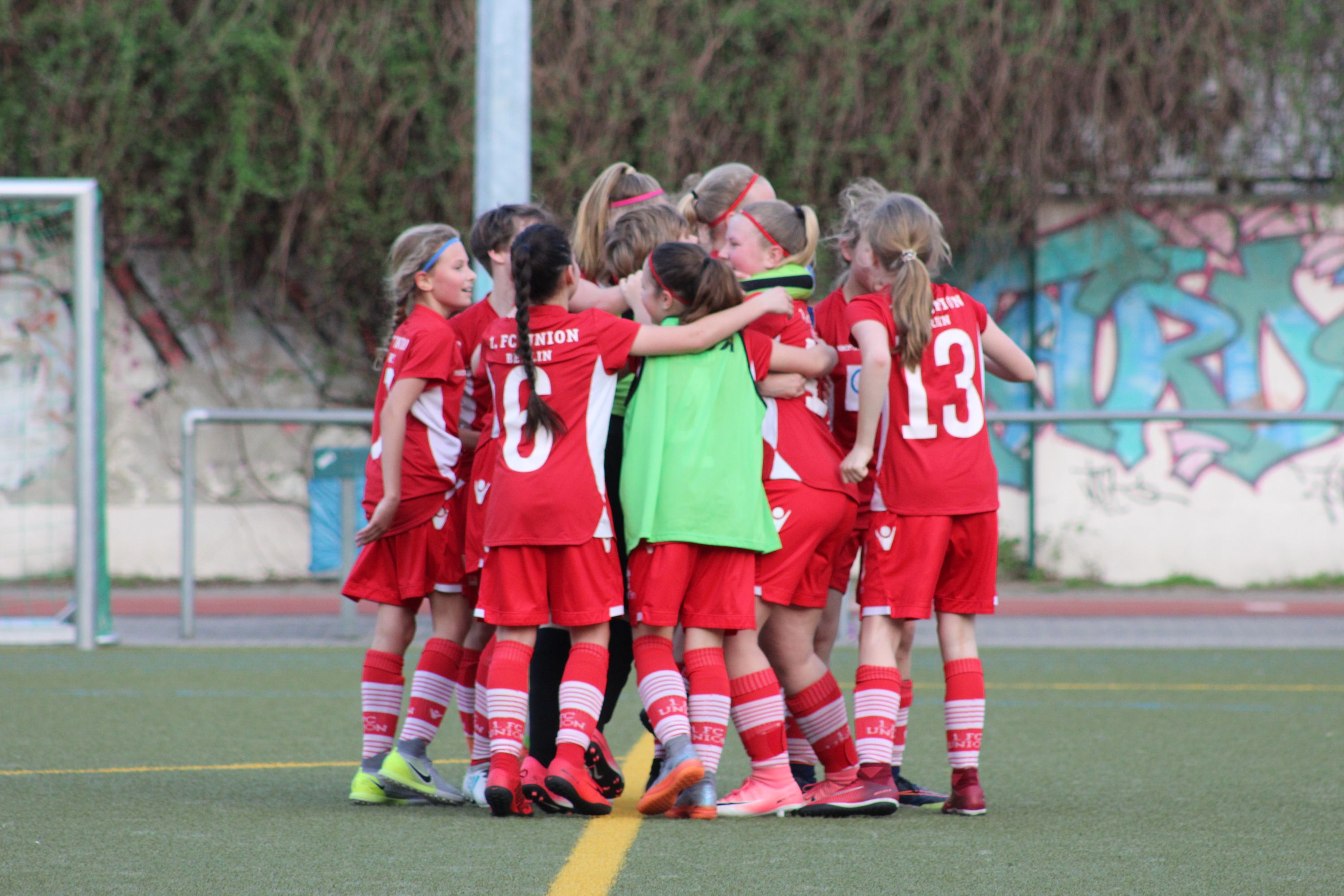 Eine Gruppe von Mädchen in roten Fußballtrikots umarmt sich jubelnd auf einem Sportplatz.