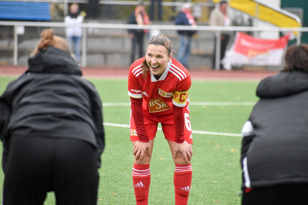 Lächelnde Fußballspielerin in rotem Trikot mit Kapitänsbinde, umgeben von zwei anderen Personen, die auf einem Platz stehen.
