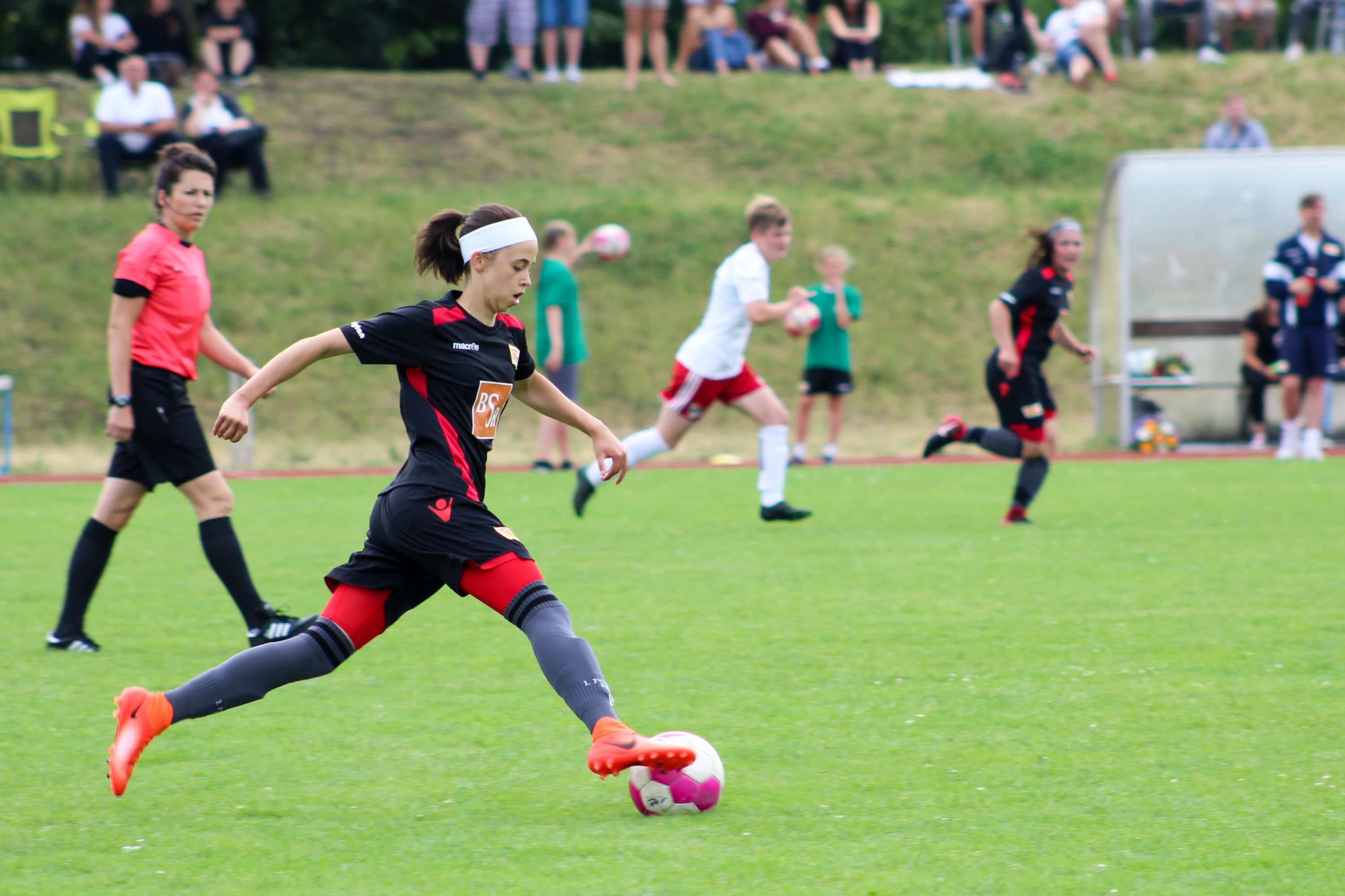 Eine spielende Fußballerin in Schwarz-Rot schießt den Ball auf einem grünen Rasenplatz, Zuschauer im Hintergrund.
