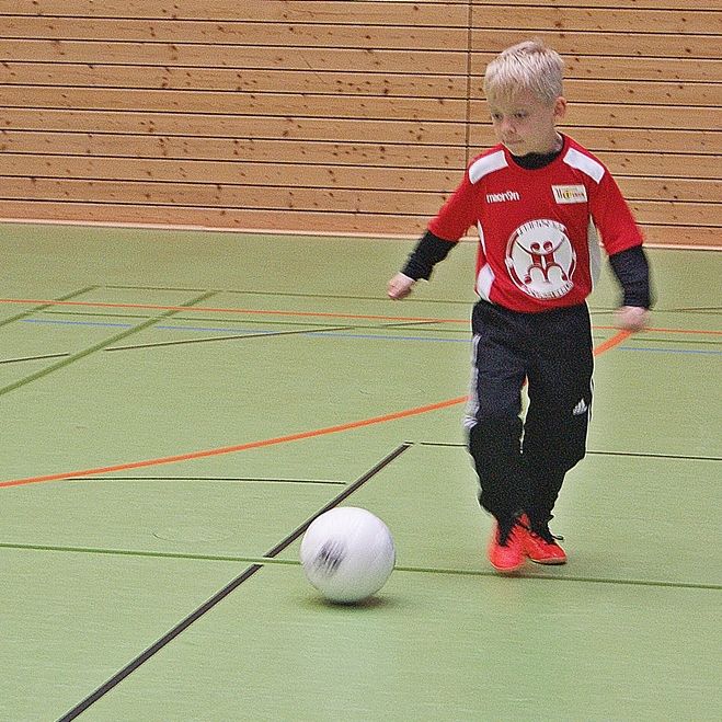 Ein Junge in einem roten Fußballtrikot dribbelt einen weißen Ball in einer Sporthalle mit grünen Bodenmarkierungen.