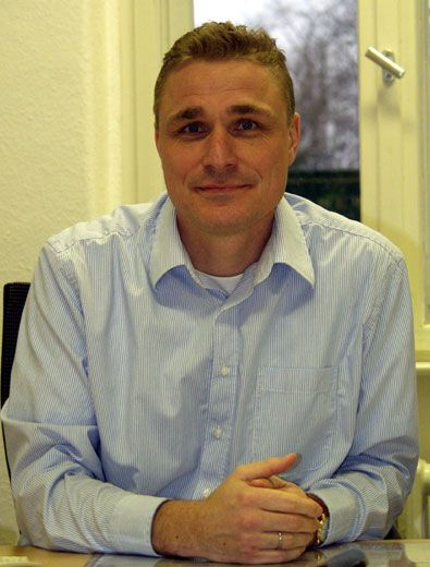 Ein lächelnder Mann in einem gestreiften Hemd sitzt an einem Tisch vor einem Fenster.