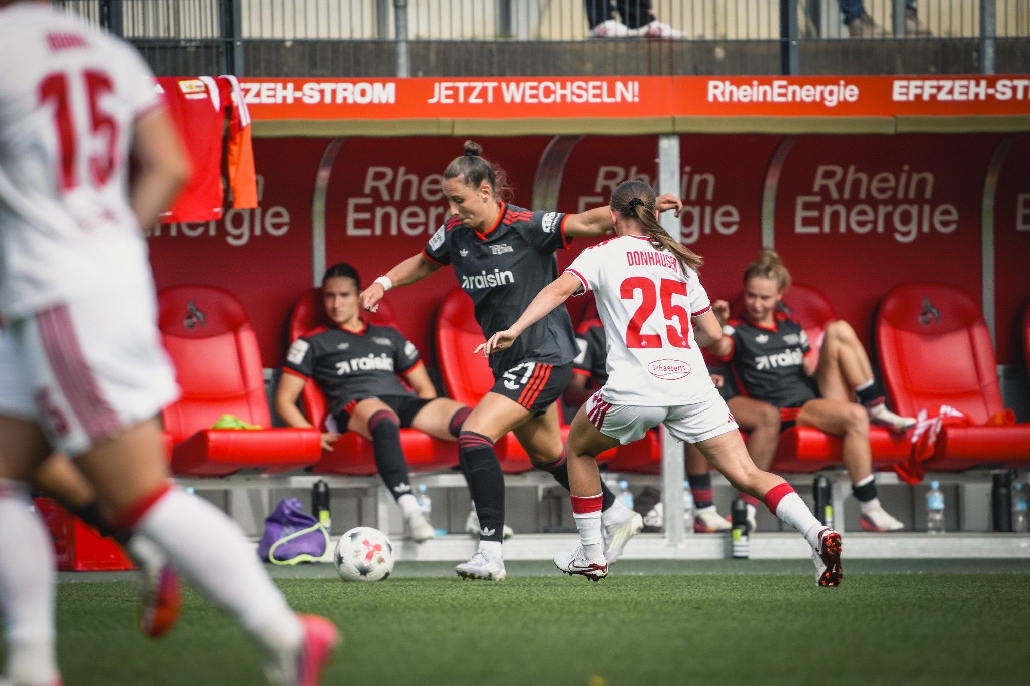 Eine Fußballspielerin in schwarz dribbelt den Ball, während eine Spielerin in weiß versucht, den Ball zu erobern. Hintergrund: Bank mit Spielenden.