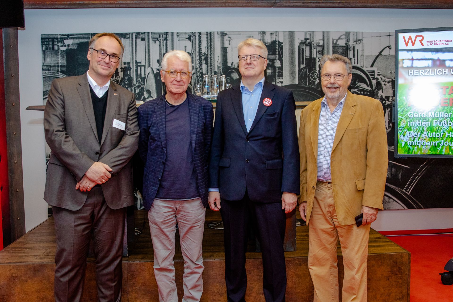 Vier Männer in Anzug und eleganter Kleidung stehen lächelnd nebeneinander vor einer industriellen Hintergrundkulisse.