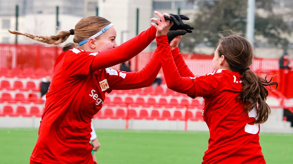 Zwei Fußballspielerinnen in roten Trikots umarmen sich freudig auf einem Spielfeld.