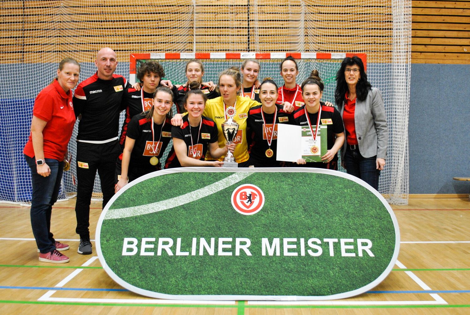 Eine Gruppenaufnahme von Sportlerinnen mit Medaillen und Pokalen, die stolz vor einem Banner "BERLINER MEISTER" stehen.
