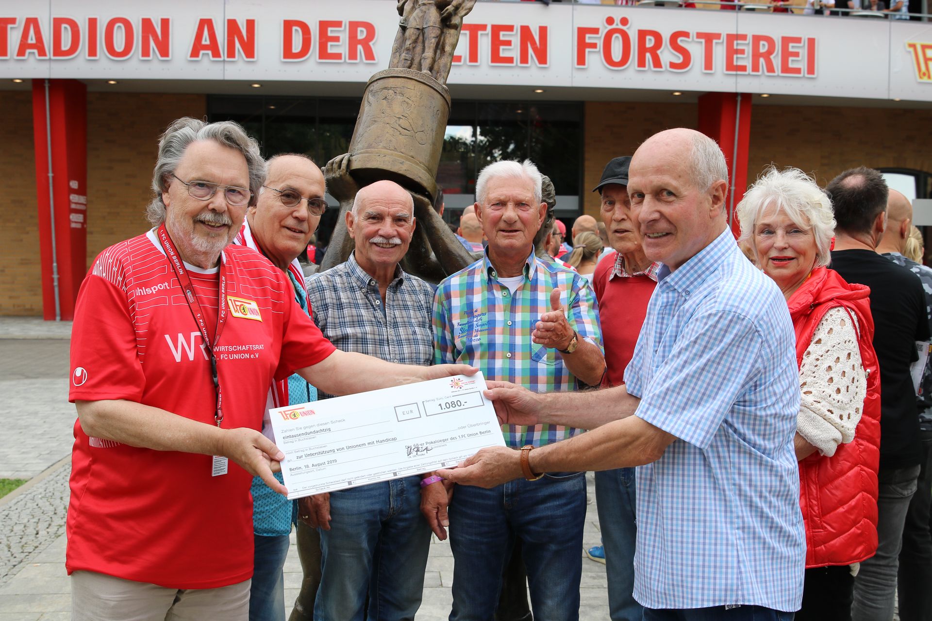 Eine Gruppe von Männern und Frauen nimmt einen Scheck entgegen, während sie vor einem Gebäude mit einer Statue stehen.