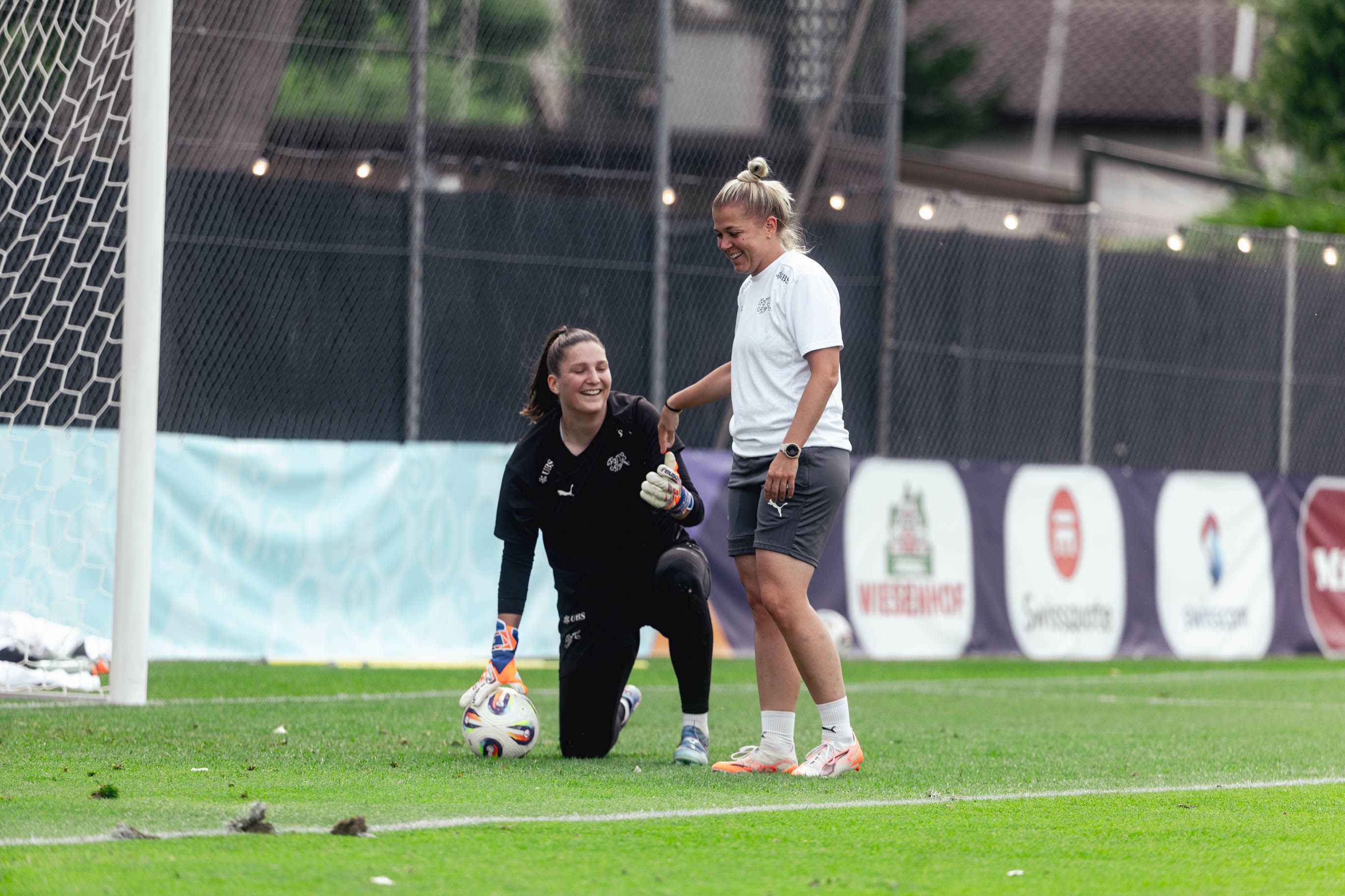 Zwei Frauen in Sportkleidung stehen auf einem Fußballfeld. Eine kniet vor dem Tor, beide lachen und haben Spaß.