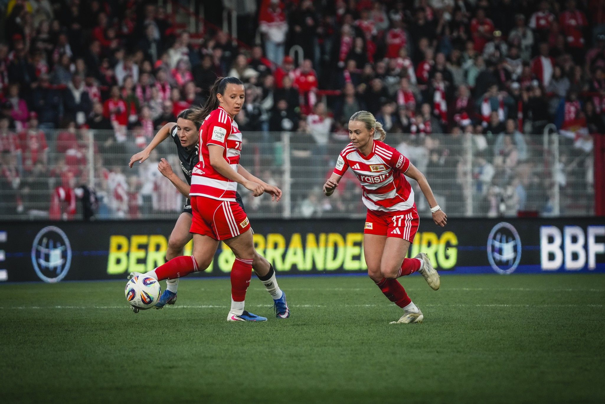 Drei Fußballspielerinnen in roten Trikots kämpfen um den Ball auf einem Spielfeld, im Hintergrund jubelnde Fans.