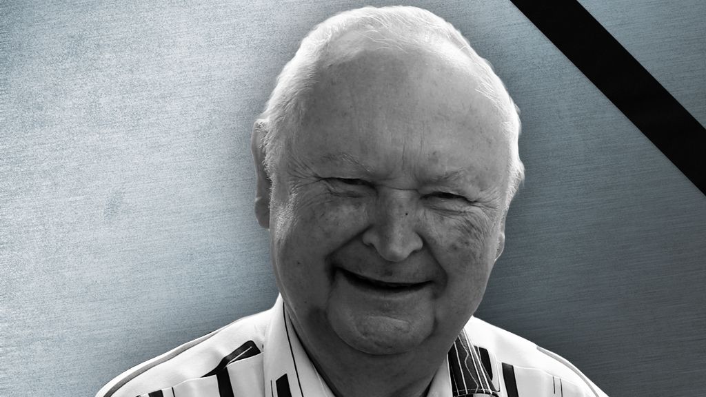 Ein lächelnder älterer Mann mit weißem Haar und gestreifter Bluse vor einem grauen Hintergrund.