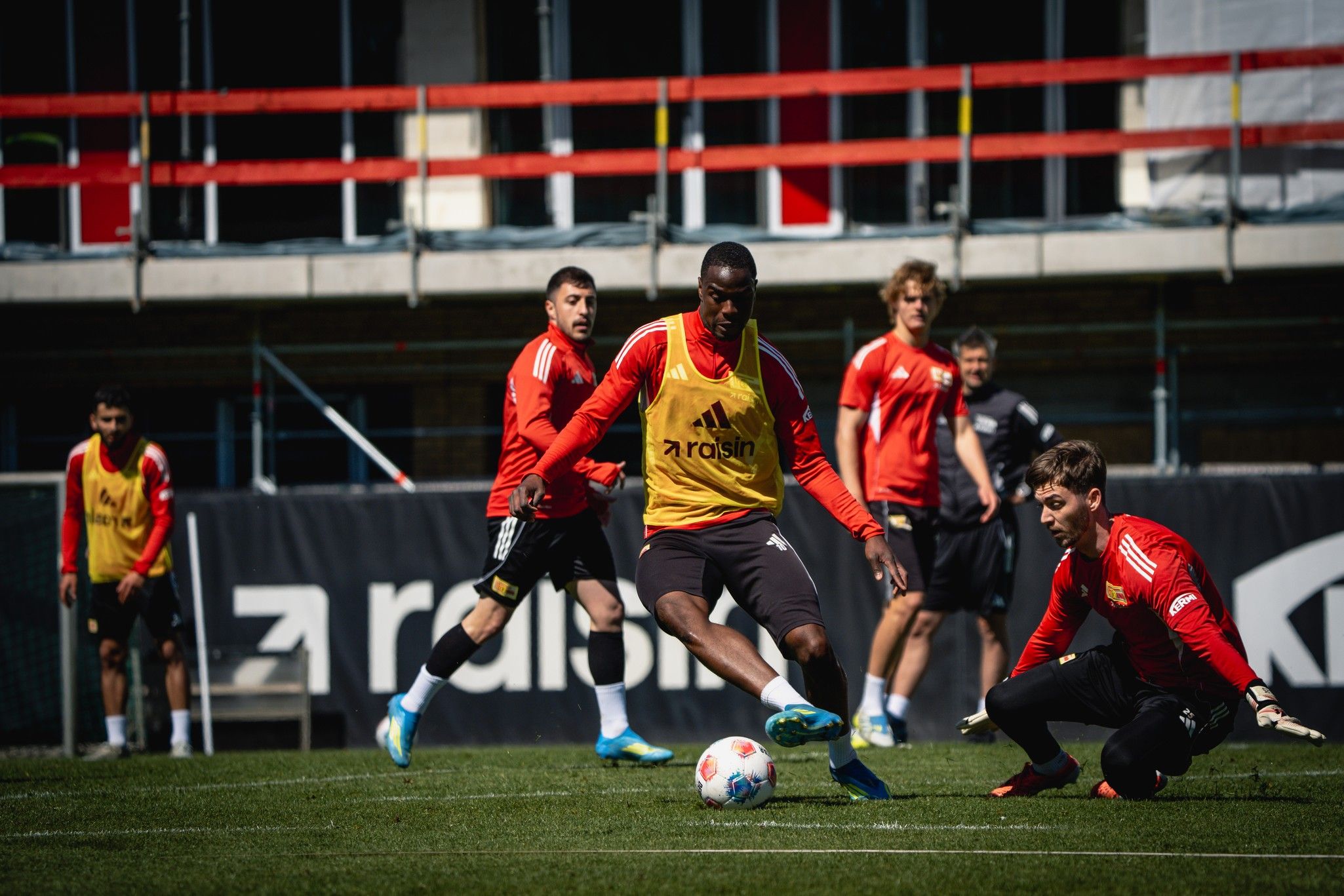 Fußballtraining mit mehreren Spielern auf dem Platz, wobei ein Spieler den Ball schießt und ein Torwart sich bereit macht.