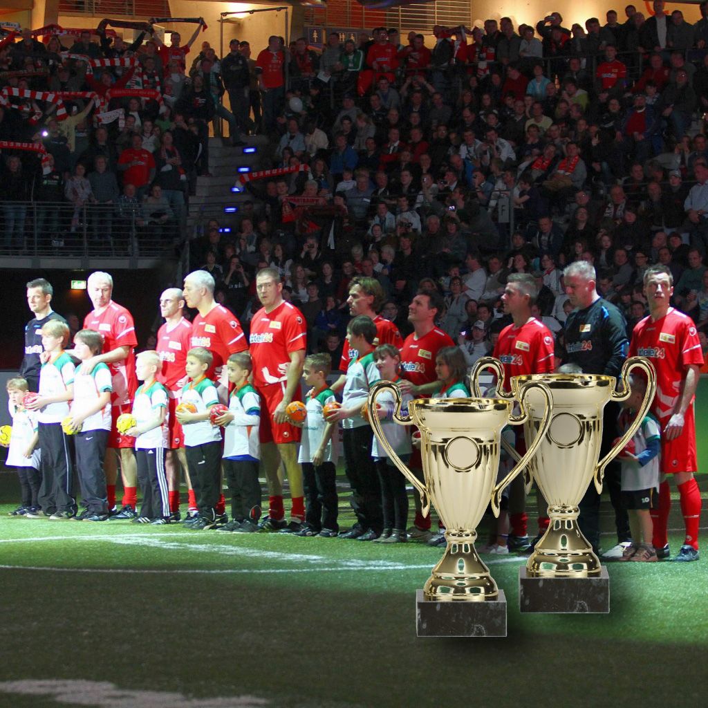 Fußballmannschaft steht auf dem Spielfeld, umgeben von Zuschauern, während zwei Pokale im Vordergrund stehen.