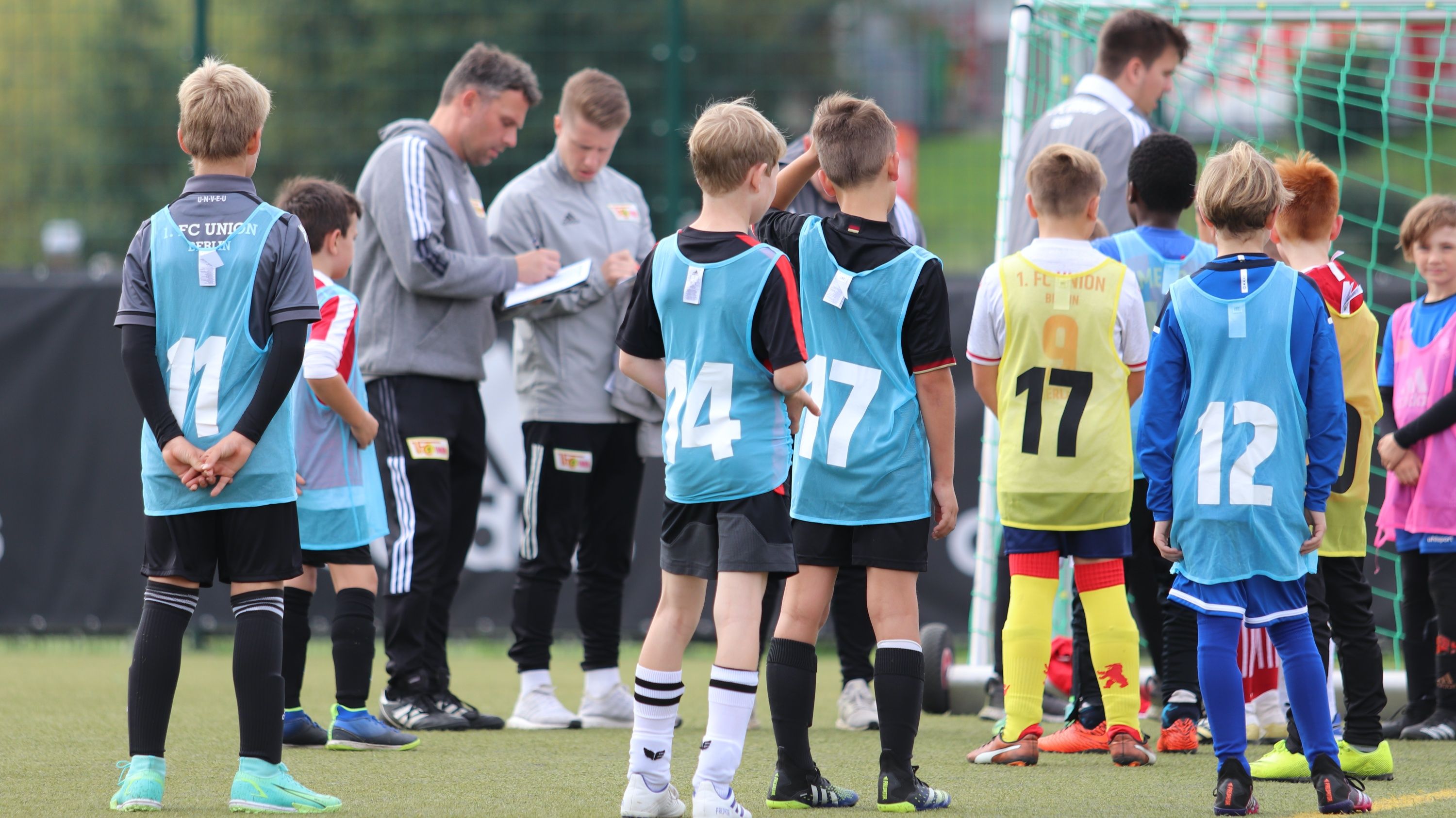 Junge Fußballspieler in blauen Westen stehen auf dem Feld, während Trainer Anweisungen gibt und andere Spieler zuschauen.