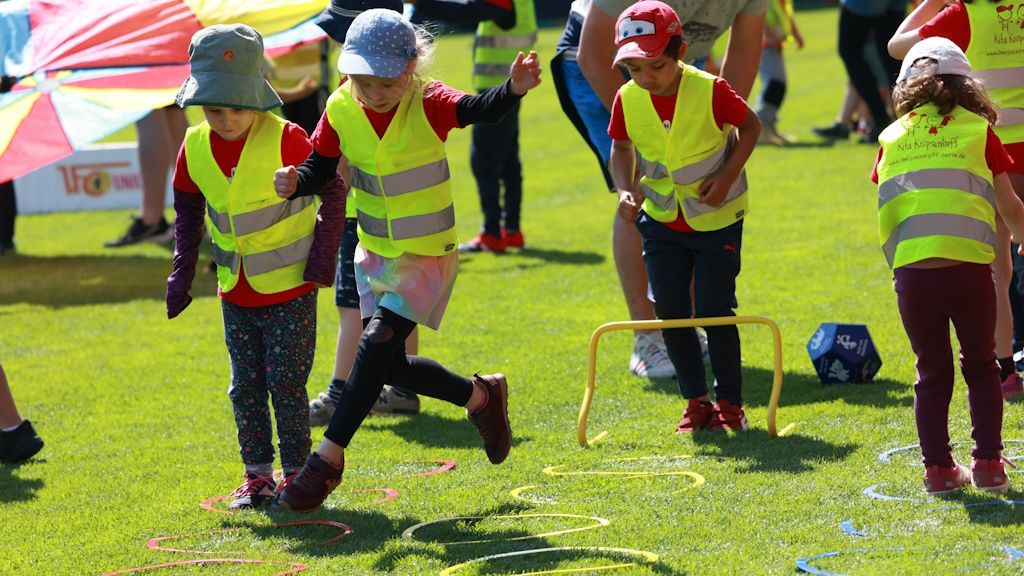 Vier Kinder in leuchtenden Sicherheitswesten springen über Hütchen auf einem grünen Rasen während eines Outdoor-Spiels.