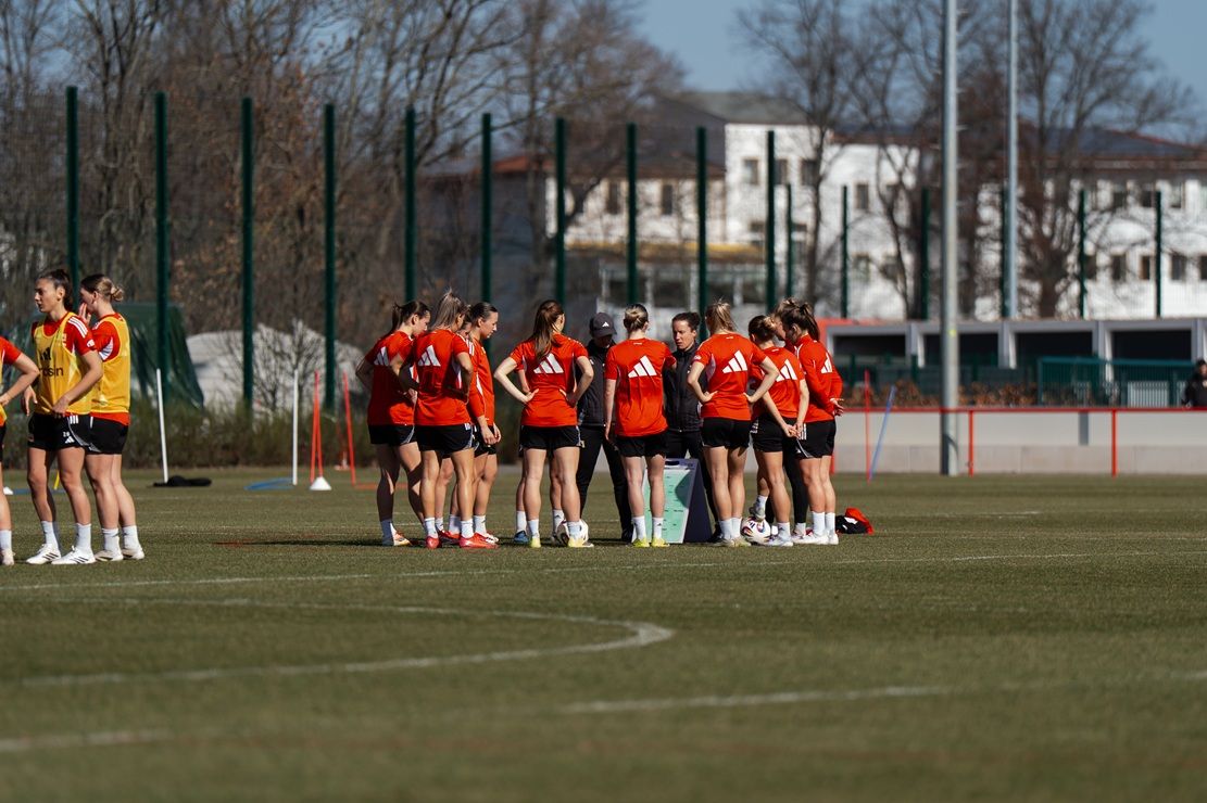 Gruppierung von Fußballspielerinnen in roten Trikots auf einem Trainingsplatz, umgeben von Bäumen und Gebäuden im Hintergrund.