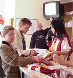 Ein Verkaufstresen mit Kunden, die Artikel kaufen, und einer Verkäuferin, die eine Bestellung entgegennimmt. Im Hintergrund ein Fernseher.