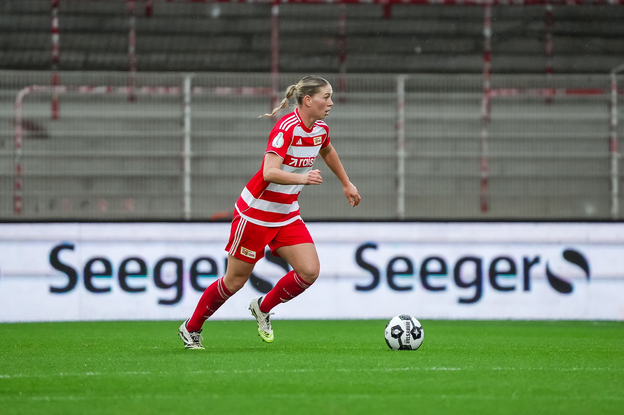 Eine Spielerin in rotem Fußballtrikot läuft über das Feld, während im Hintergrund Werbung zu sehen ist.