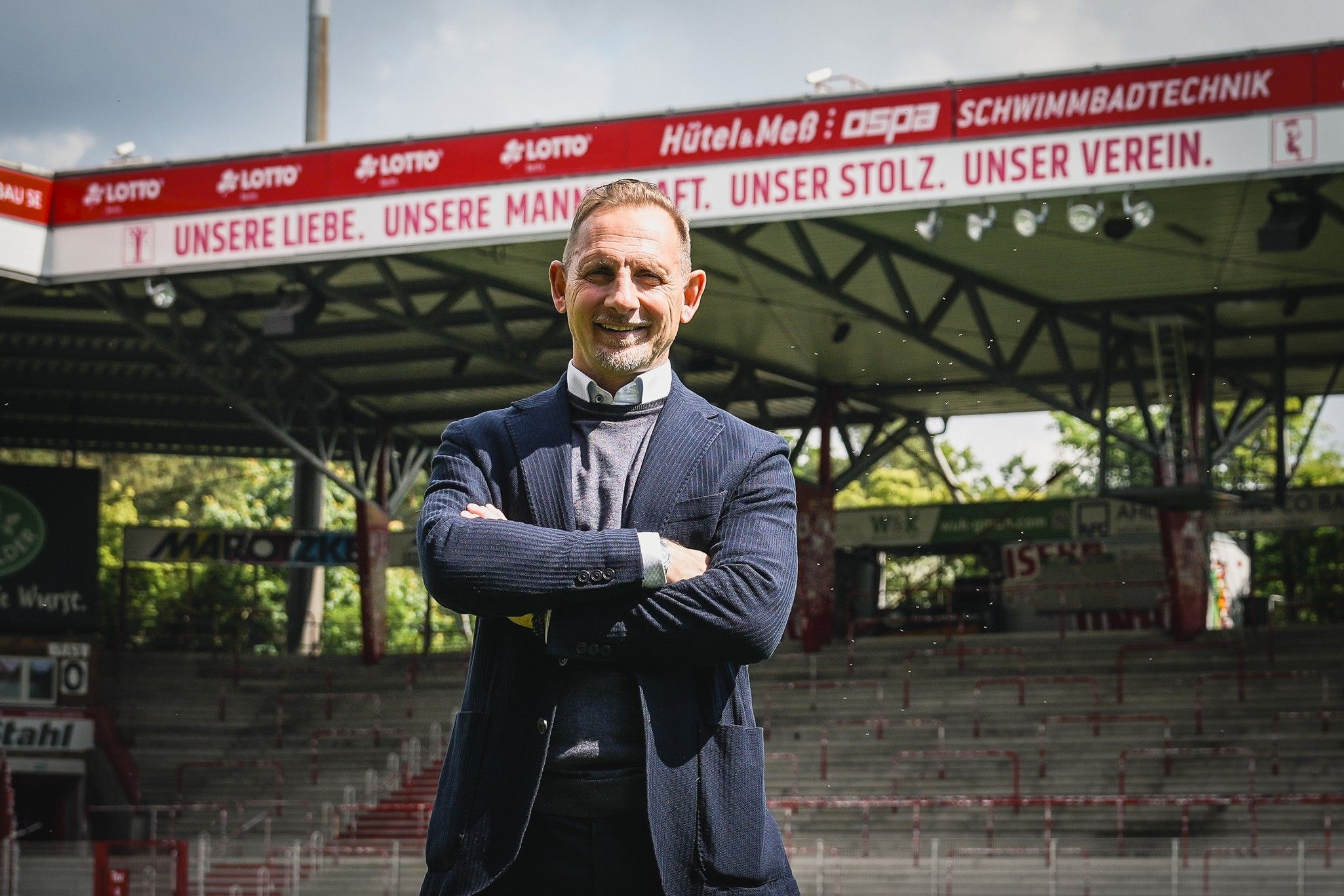 Lächelnder Mann im Anzug steht vor einer Sportstadion-Gegend mit Tribünen und Werbeschildern im Hintergrund.