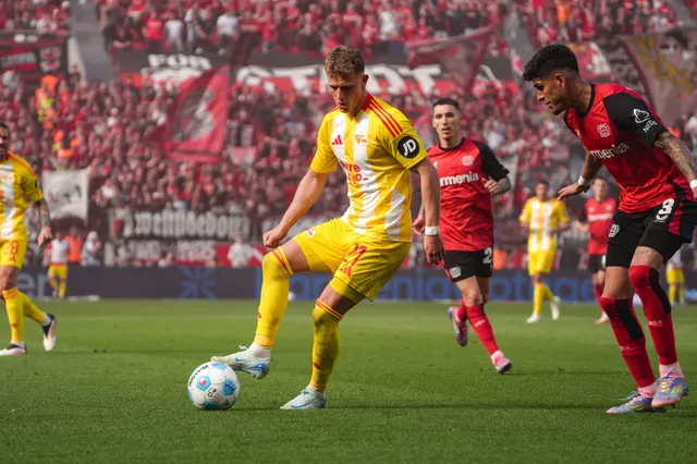 Spielszene eines Fußballspiels mit Spielern in gelben und roten Trikots auf einem grünen Rasenfeld, Fans im Hintergrund.