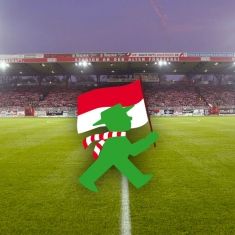 Ein grüner Comic-Charakter mit einer Flagge läuft über ein Fußballfeld, umgeben von Stadionlicht und Zuschauertribünen.