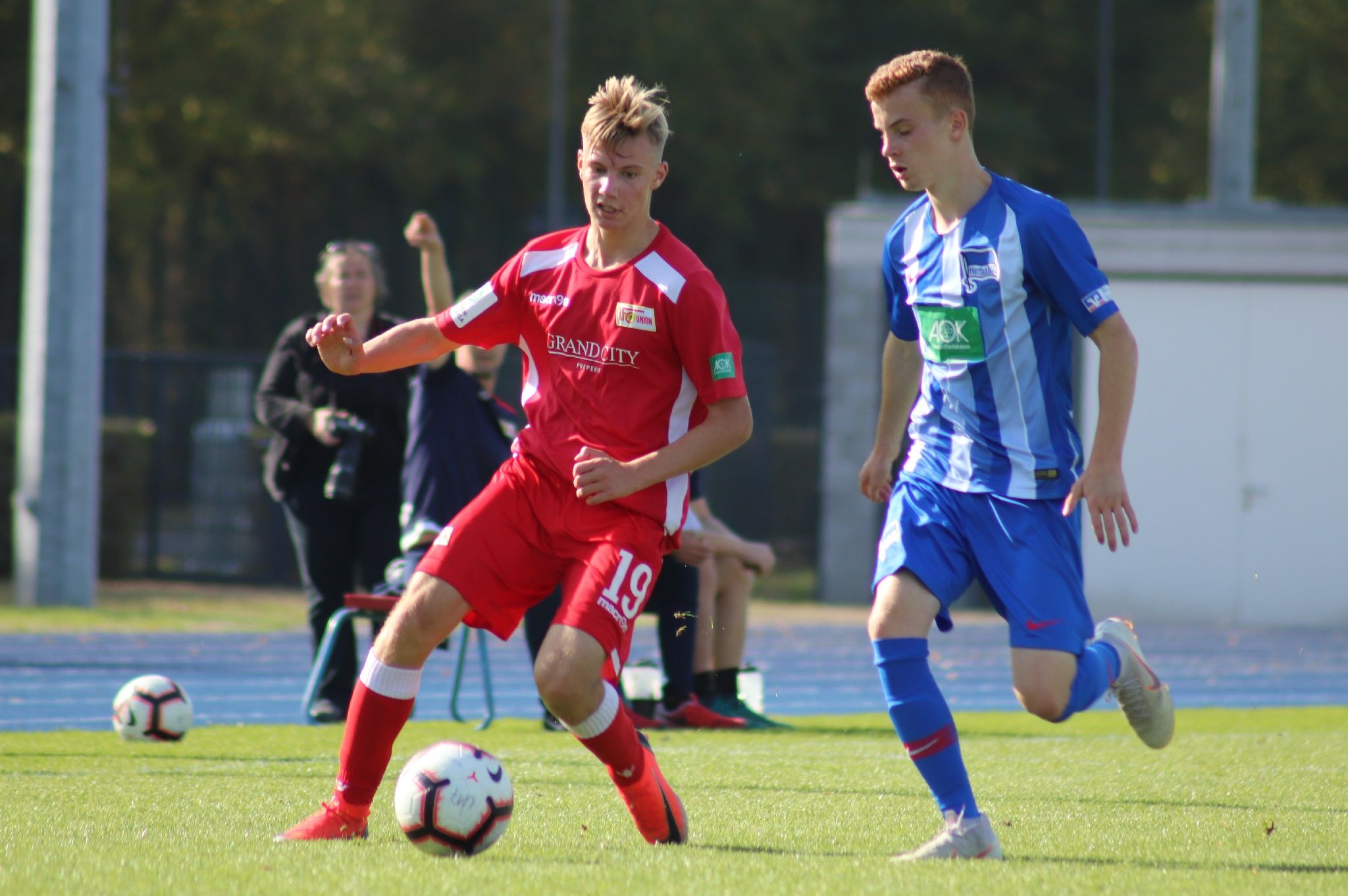 Ein Fußballspiel zwischen zwei Spielern, einer in rotem und einer in blauem Trikot, auf einem grünen Rasenplatz.