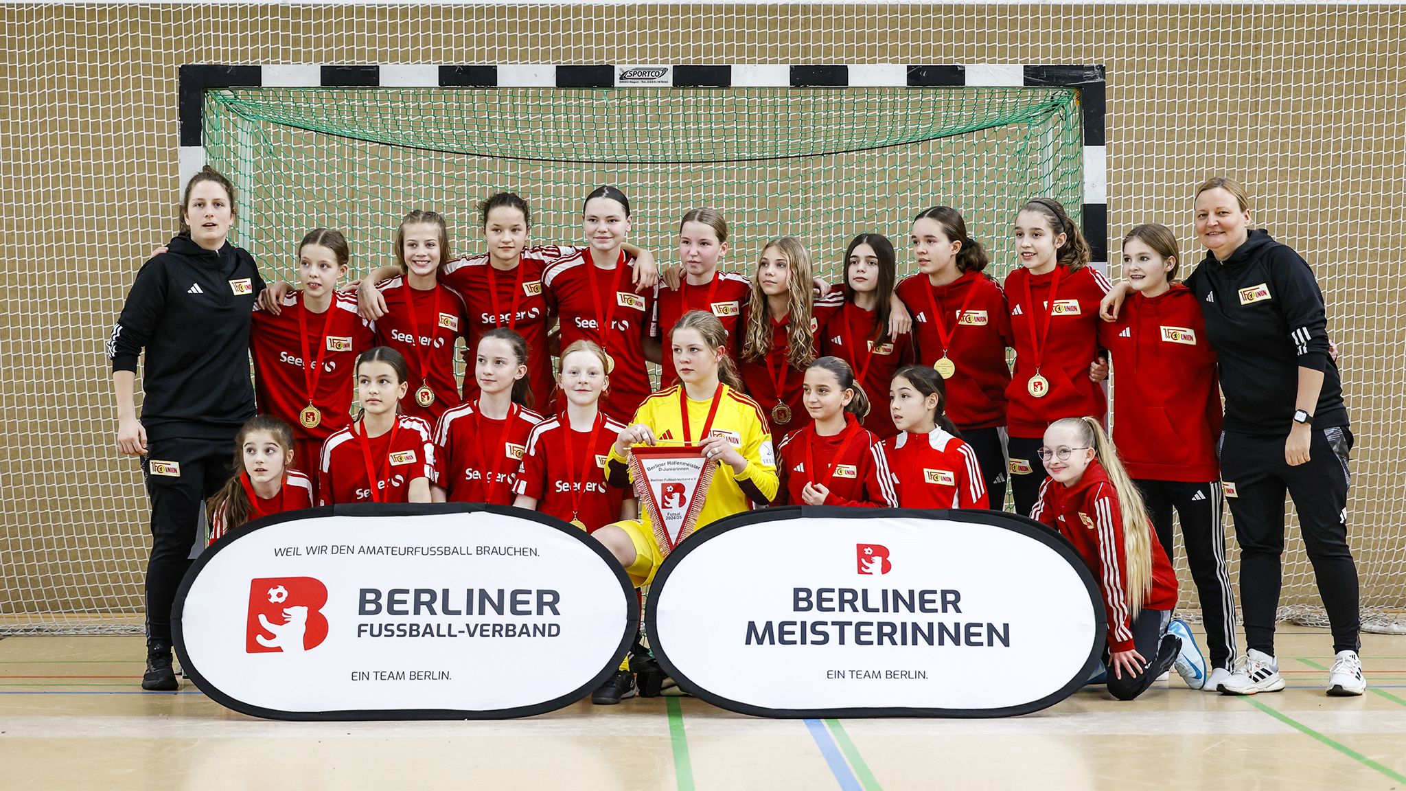 Gruppe von Mädchen in roten Trikots, die nach einem Handballspiel lächeln, zusammen mit Medaillen und Pokal vor einem Tor.
