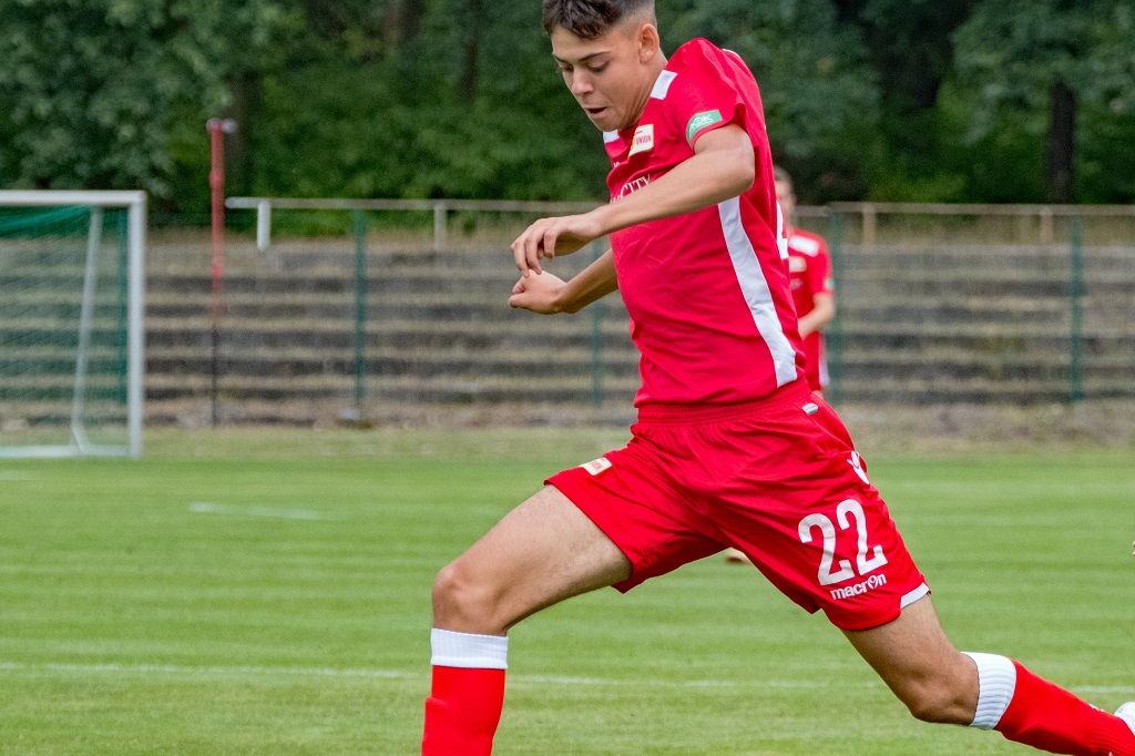 Fußballspieler in rotem Trikot mit der Nummer 22, der in einer Wiese läuft und den Ball spielt.