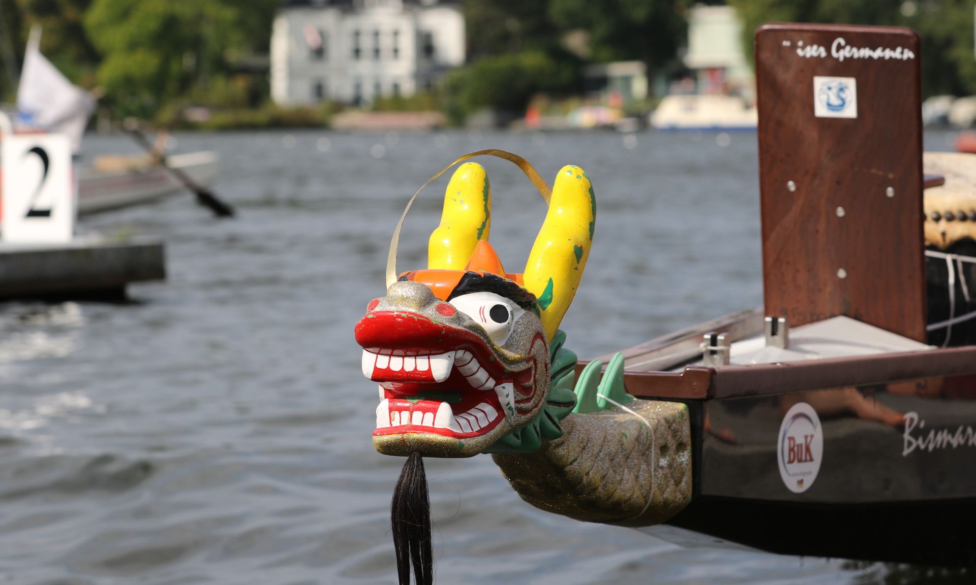 Drachenbootkopf mit buntem Drachenmotiv, am Ufer eines Gewässers mit Booten im Hintergrund.