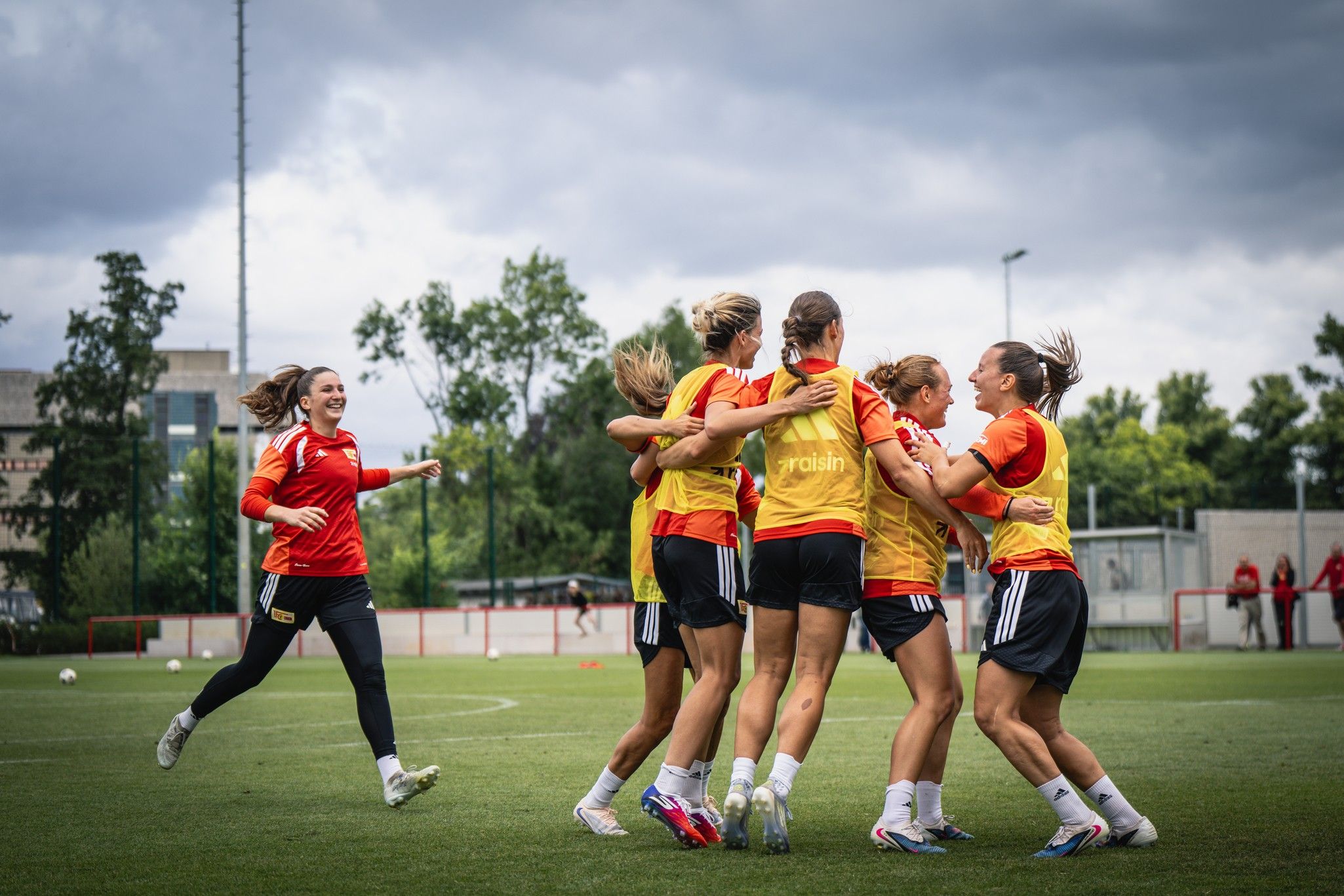 Sechs Frauen in roten Trikots umarmen sich fröhlich auf einem Fußballplatz unter bewölkten Himmel.