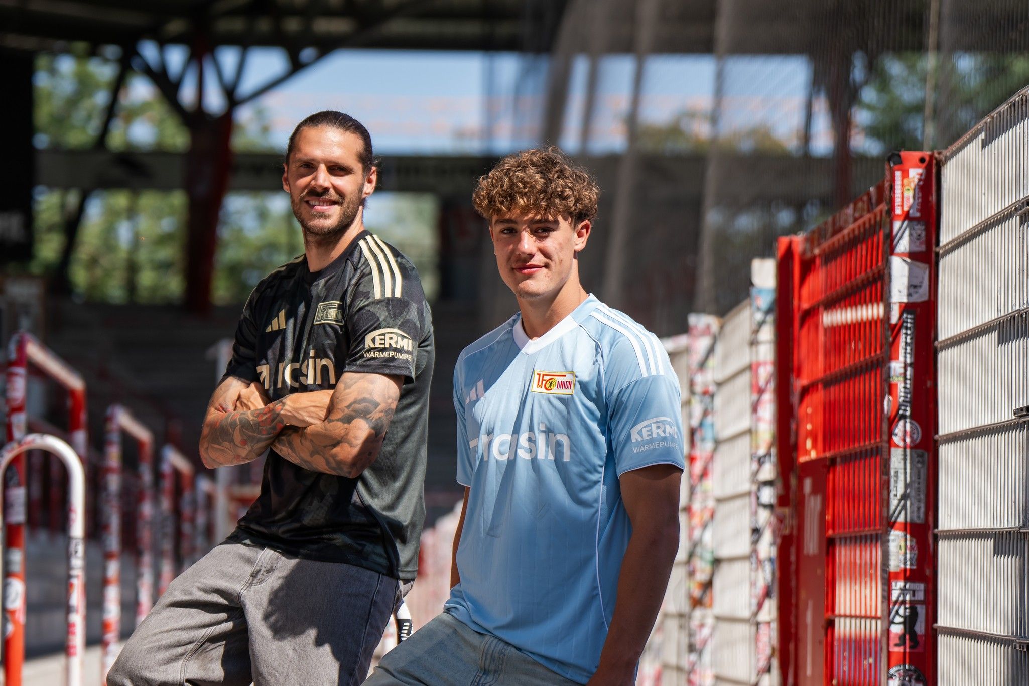 Zwei junge Männer stehen in Sporttrikots in einem Stadion. Der eine trägt ein schwarzes Trikot, der andere ein hellblaues.