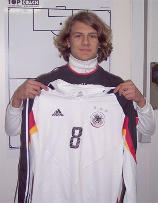 Ein junger Mann mit lockigem Haar hält ein weißes Trikot mit der Nummer 8 und dem deutschen Fußballverbandssymbol in den Händen.