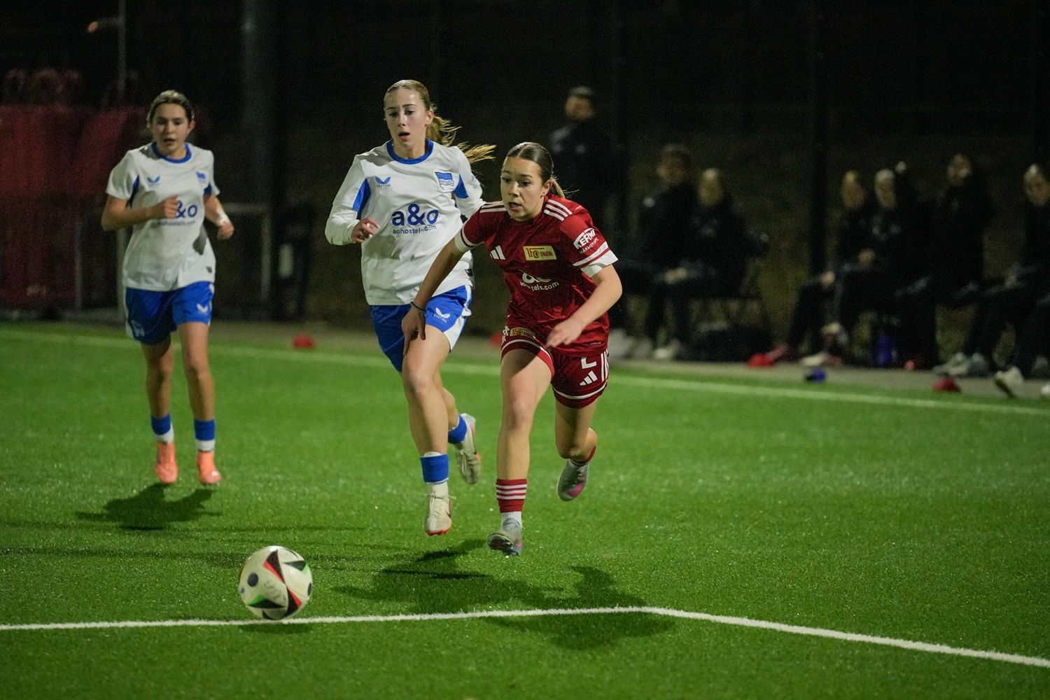 Zwei Fußballerinnen in blauen Trikots verfolgen eine Spielerin in rotem Trikot, die einen Ball auf einem Kunstrasenplatz dribbelt.