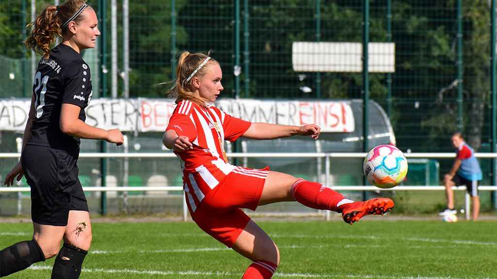 Eine Fußballspielerin in rotem Trikot mit weißen Streifen kickt den Ball, während eine gegnerische Spielerin in Schwarz zusieht.