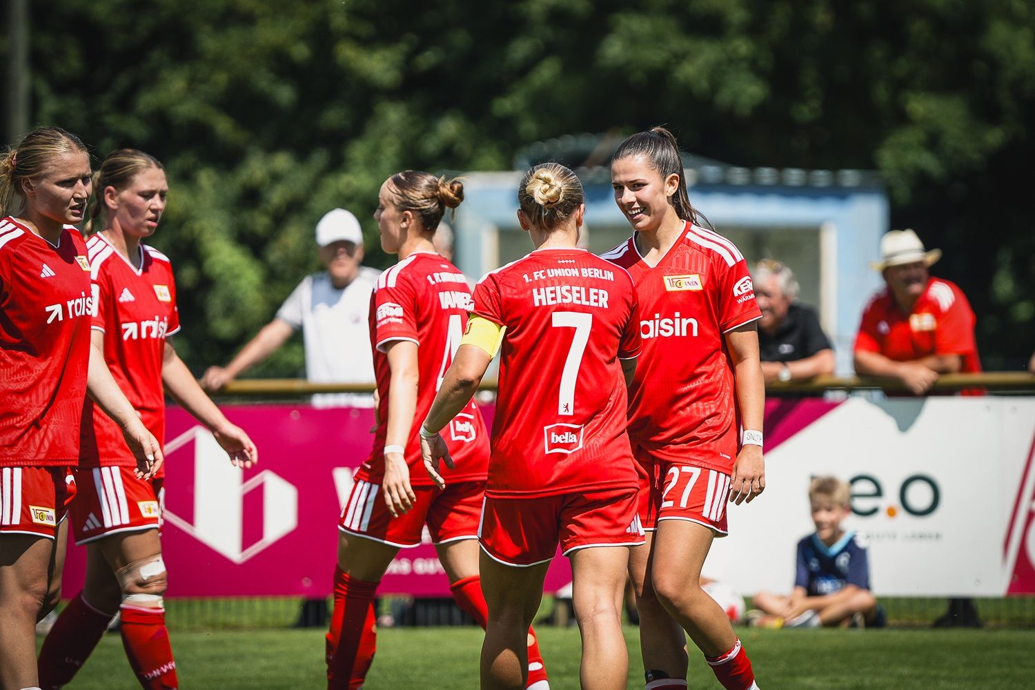 Frauenfußballmannschaft in roten Trikots feiert ein Tor, einige Spielerinnen umarmen sich auf dem Spielfeld.