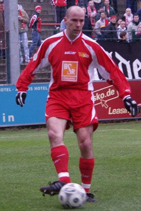 Fußballspieler in rotem Trikot und schwarzer Shorts, der mit einem Ball auf dem Platz steht.