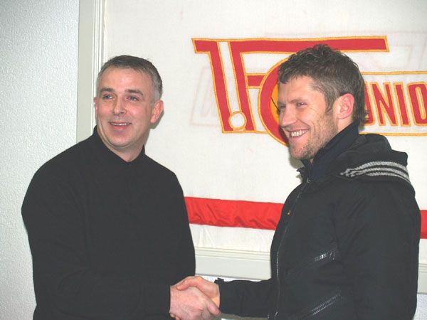 Zwei Männer schütteln sich die Hände vor einem Banner mit dem Logo eines Sportvereins im Hintergrund.