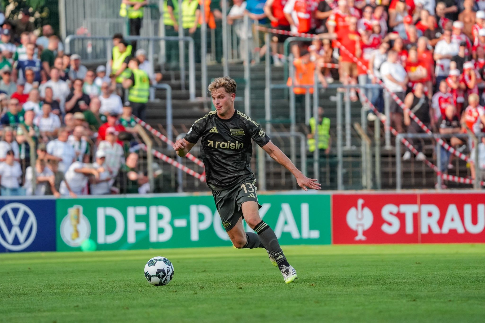 Ein Fußballspieler in einem dunklen Trikot läuft mit dem Ball auf einem Rasenfeld, während die Zuschauer im Hintergrund jubeln.