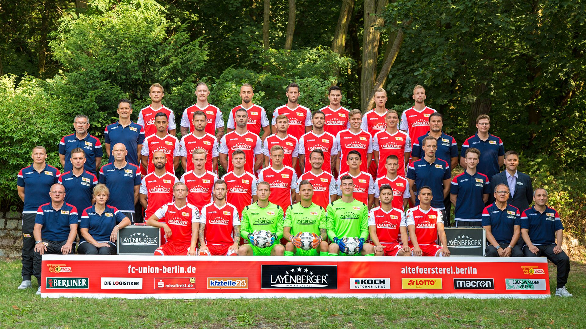 Mannschaftsfoto des FC Union Berlin mit Spielern und Trainern in roten Trikots vor einem grünen Waldbereich.