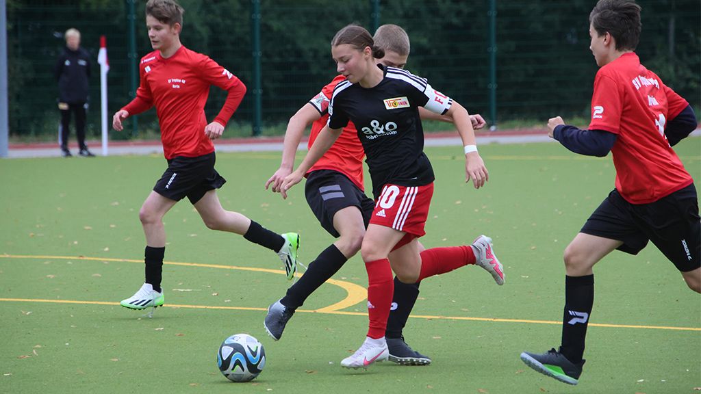 Mädchen mit Ball in Schwarz-Rot spielt Fussball auf einem Kunstrasenplatz, umgeben von spielenden Jungen in roten und schwarzen Trikots.