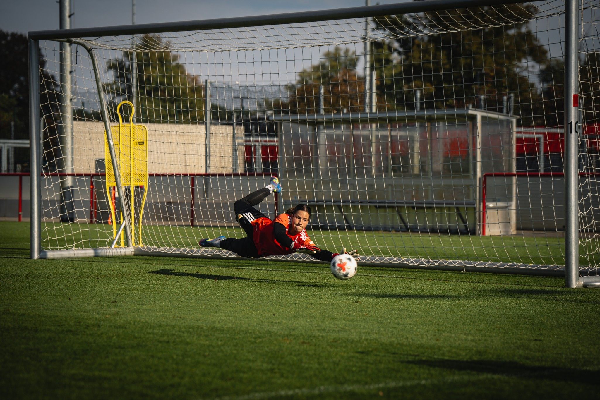 Ein Torwart springt in eine Ecke, um einen Ball im Fußballtor zu parieren, während das Training auf einem Rasenplatz stattfindet.