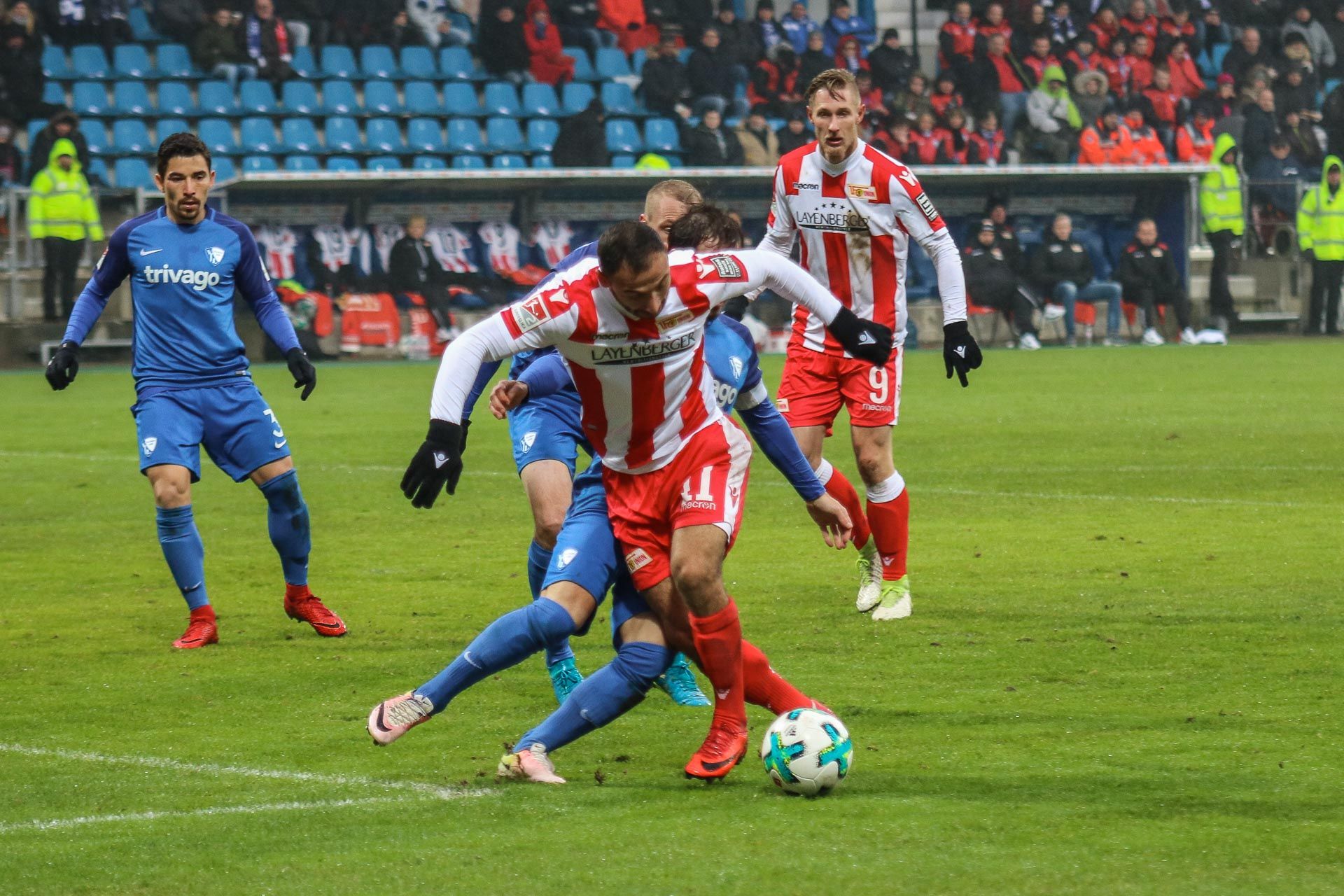 Zwei Fußballspieler kämpfen um den Ball. Sie sind auf dem Spielfeld. Im Hintergrund stehen viele Zuschauer. Sie sitzen auf der Tribüne.