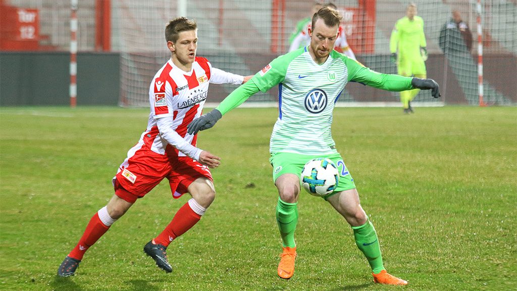 Zwei Fußballspieler in Aktion auf dem Spielfeld, einer trägt ein rotes und weißes Trikot, der andere ein grünes Trikot.