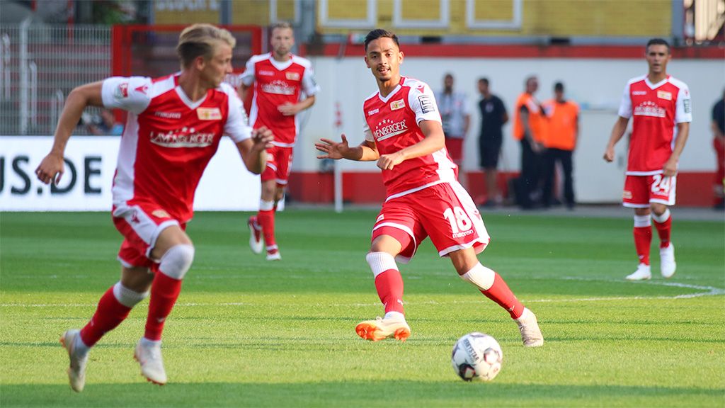 Zwei Spieler sind auf dem Rasen. Sie tragen rote Trikots. Sie spielen Fußball.