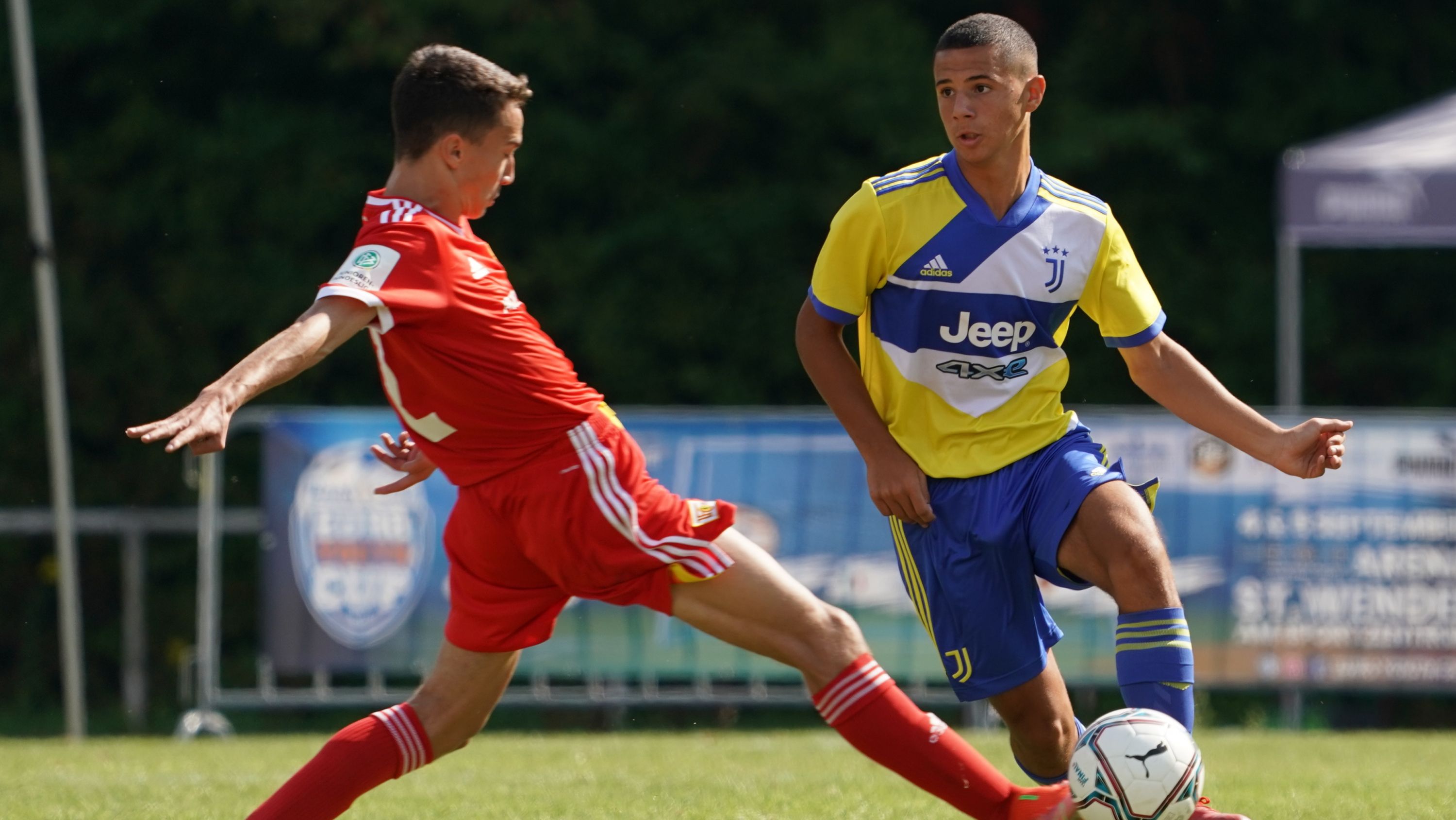 Zwei Fußballspieler in actionreichen Posen. Einer in rotem Trikot greift an, während der andere in gelbem und blauem Trikot ihm ausweicht.