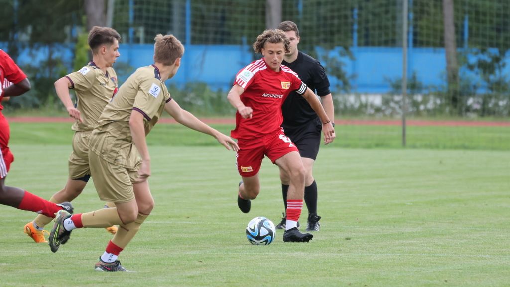 Ein Fußballspieler in roter Ausrüstung dribbelt den Ball, während zwei Gegner in beigen Trikots versuchen, ihn zu stoppen.