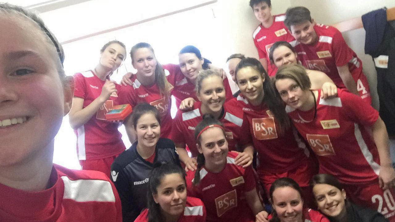 Eine Gruppe von 15 Frauen in roten Fußballtrikots posiert lächelnd für ein Selfie in einem Umkleideraum.